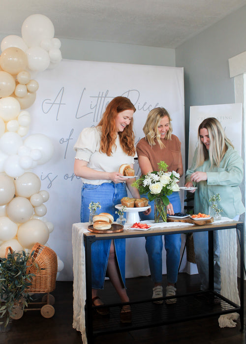 Two women standing behind a table with food and drinks, surrounded by balloons and "A Little Piece of Heaven" backdrop.