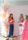 Three women at a baby shower with a balloon arch and pink backdrop.