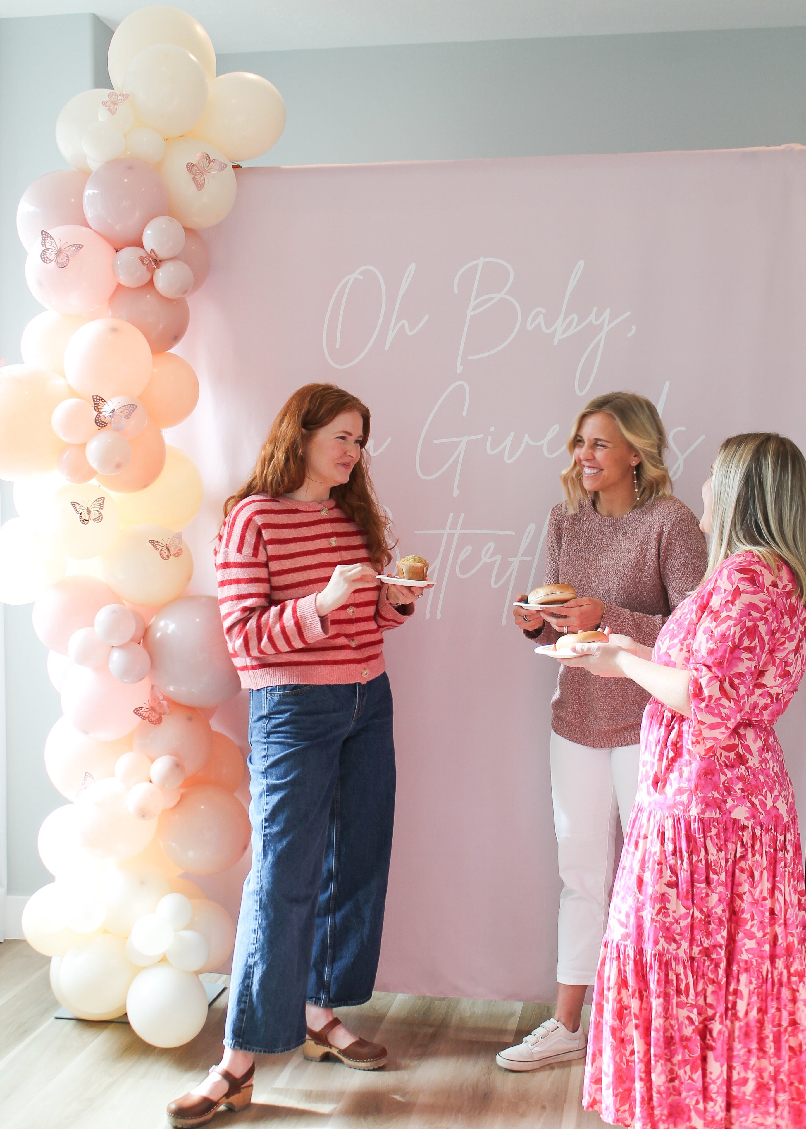 Three women at a baby shower with a balloon arch and pink backdrop.