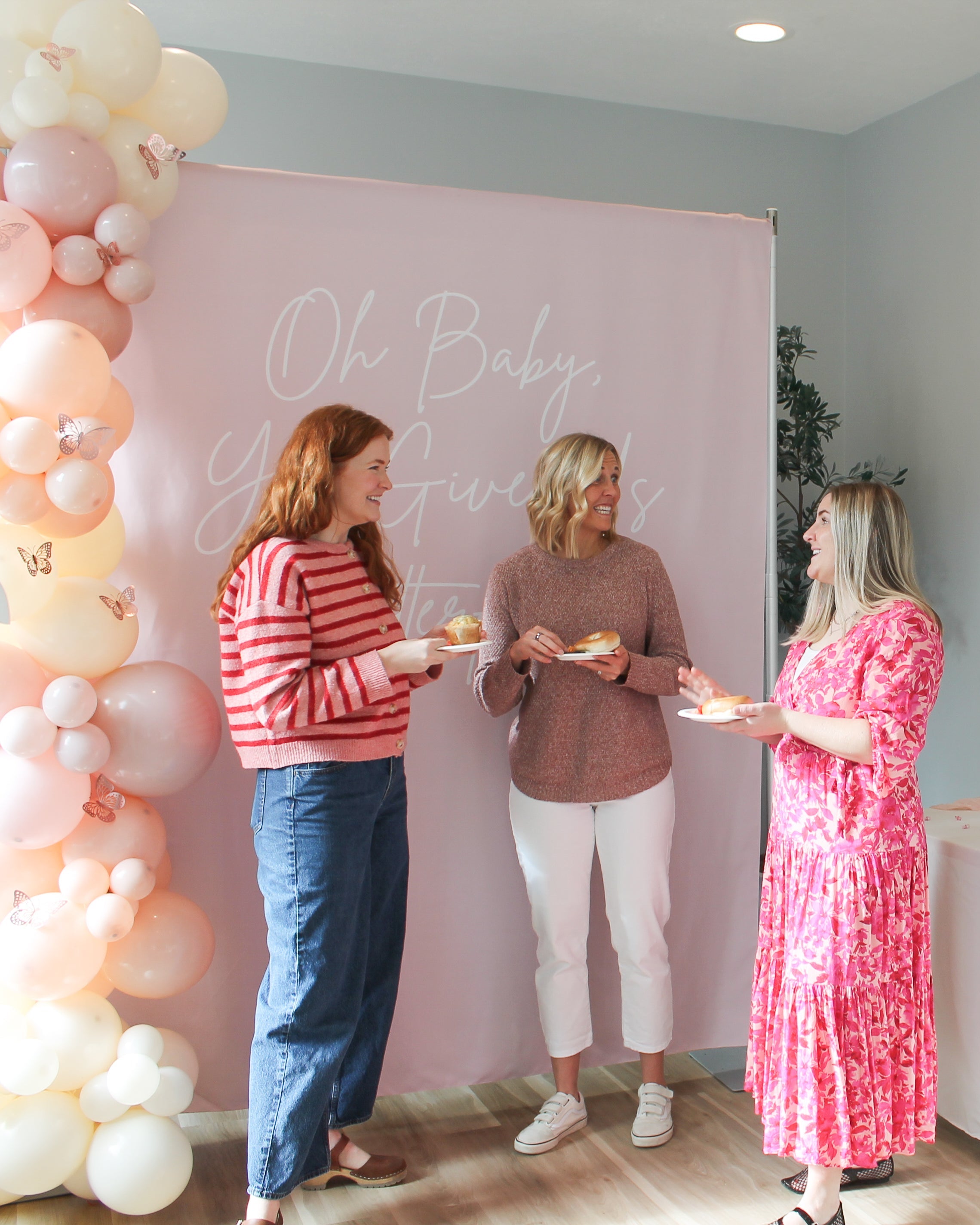 Three women at a baby shower with balloons and a pink backdrop.
