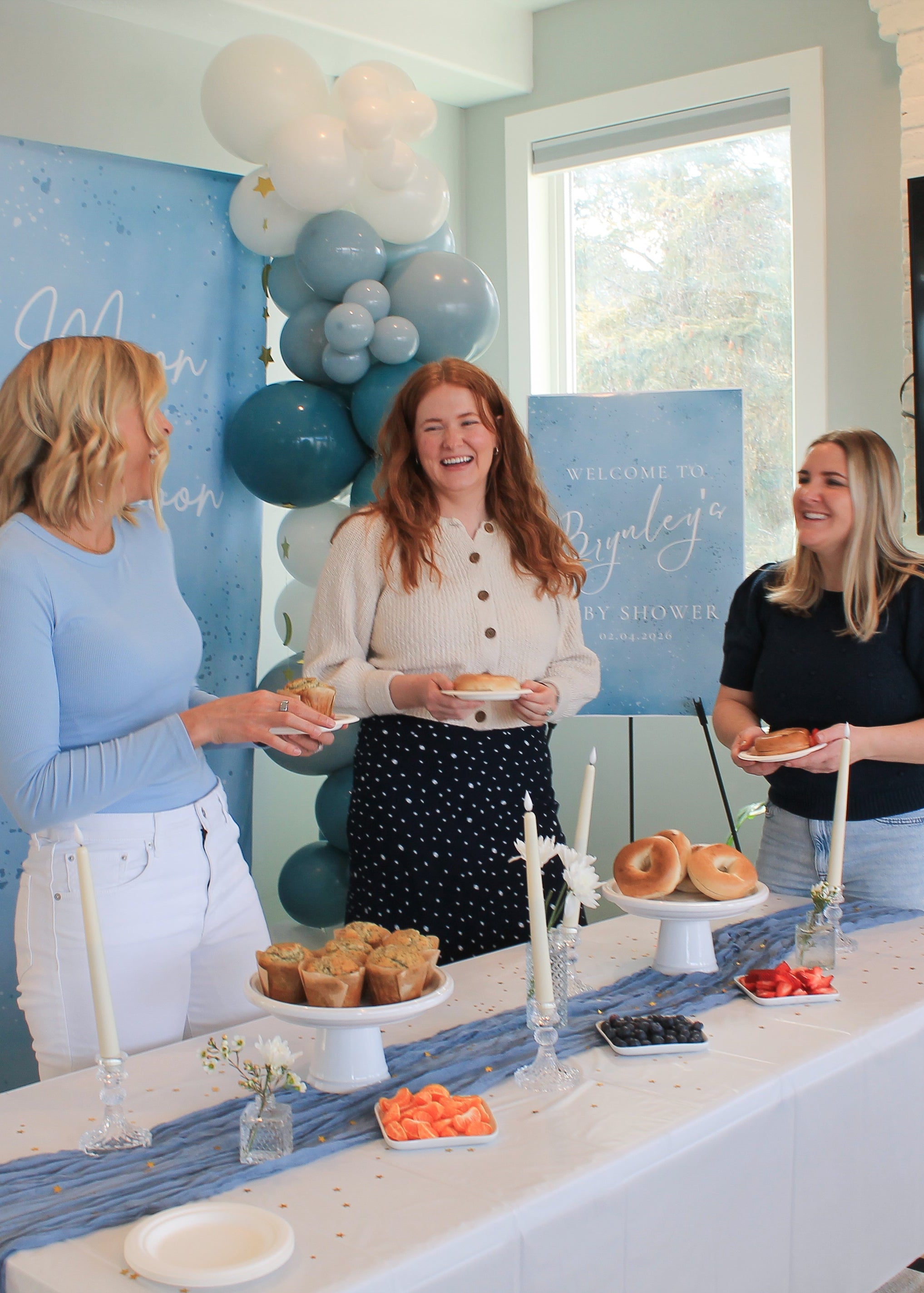 Three women standing around a table with food at a baby shower, decorated with balloons and a sign.