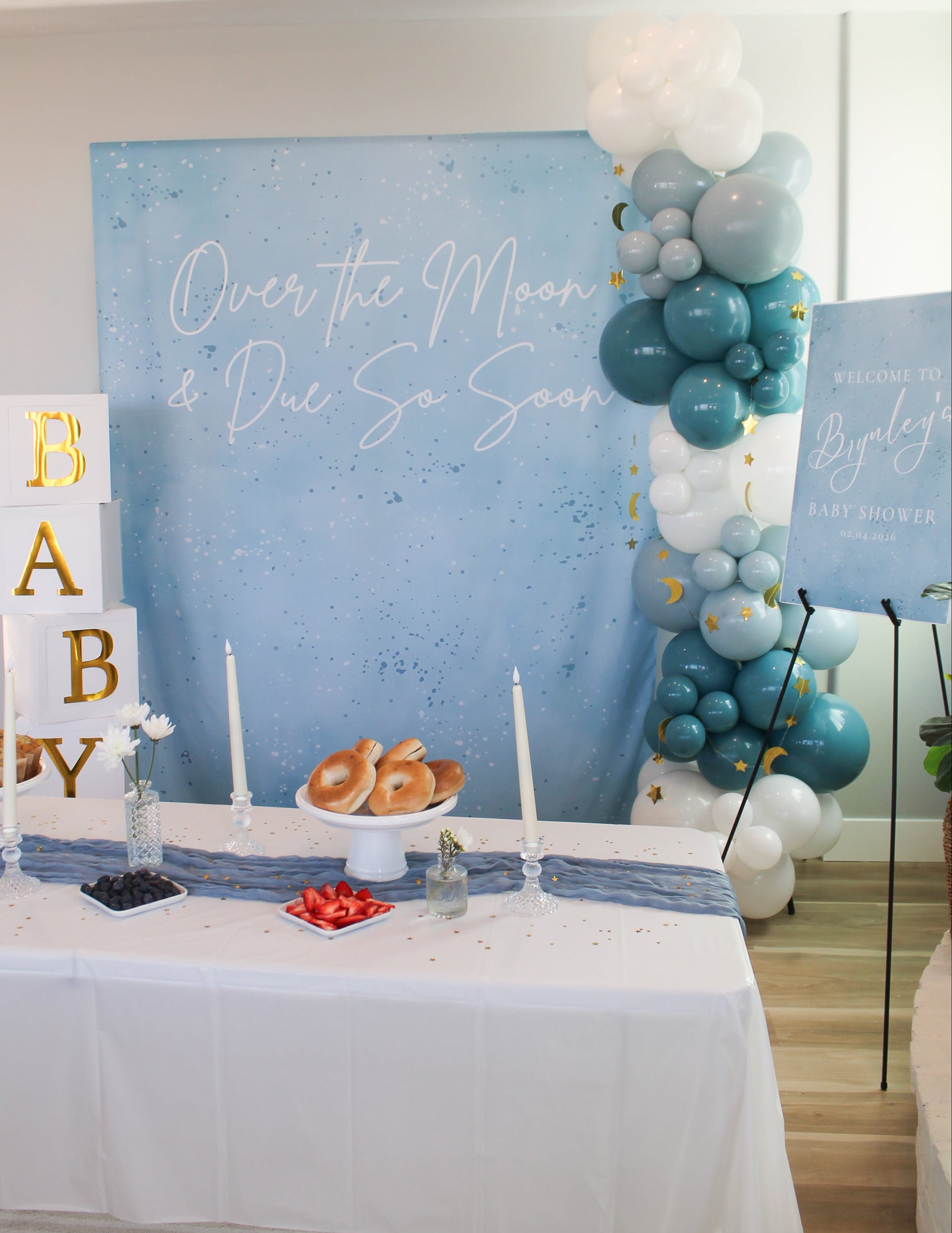 Baby shower setup with balloons, cake, and 'Baby' sign against a blue backdrop.